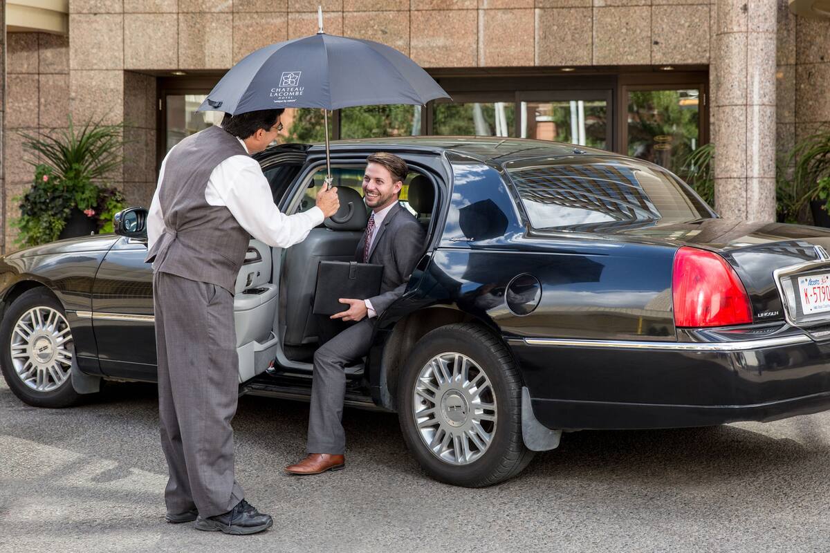 A man in a suit exiting a black luxury car with a black umbrella.