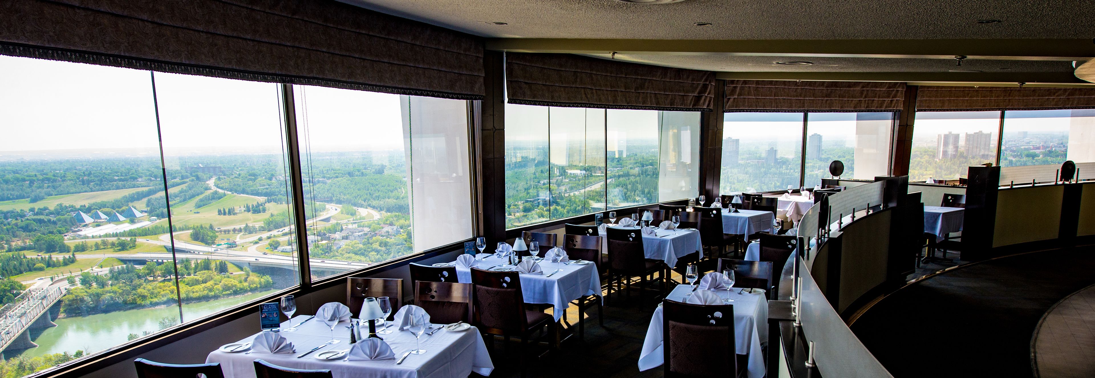 Un restaurante con mesas dispuestas para comer, grandes ventanales que ofrecen vistas a un río y al paisaje urbano.