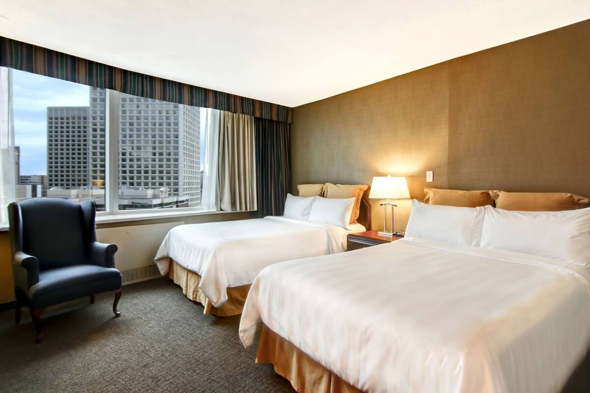 A hotel room with two beds, a chair, a nightstand, and a window with a city view.