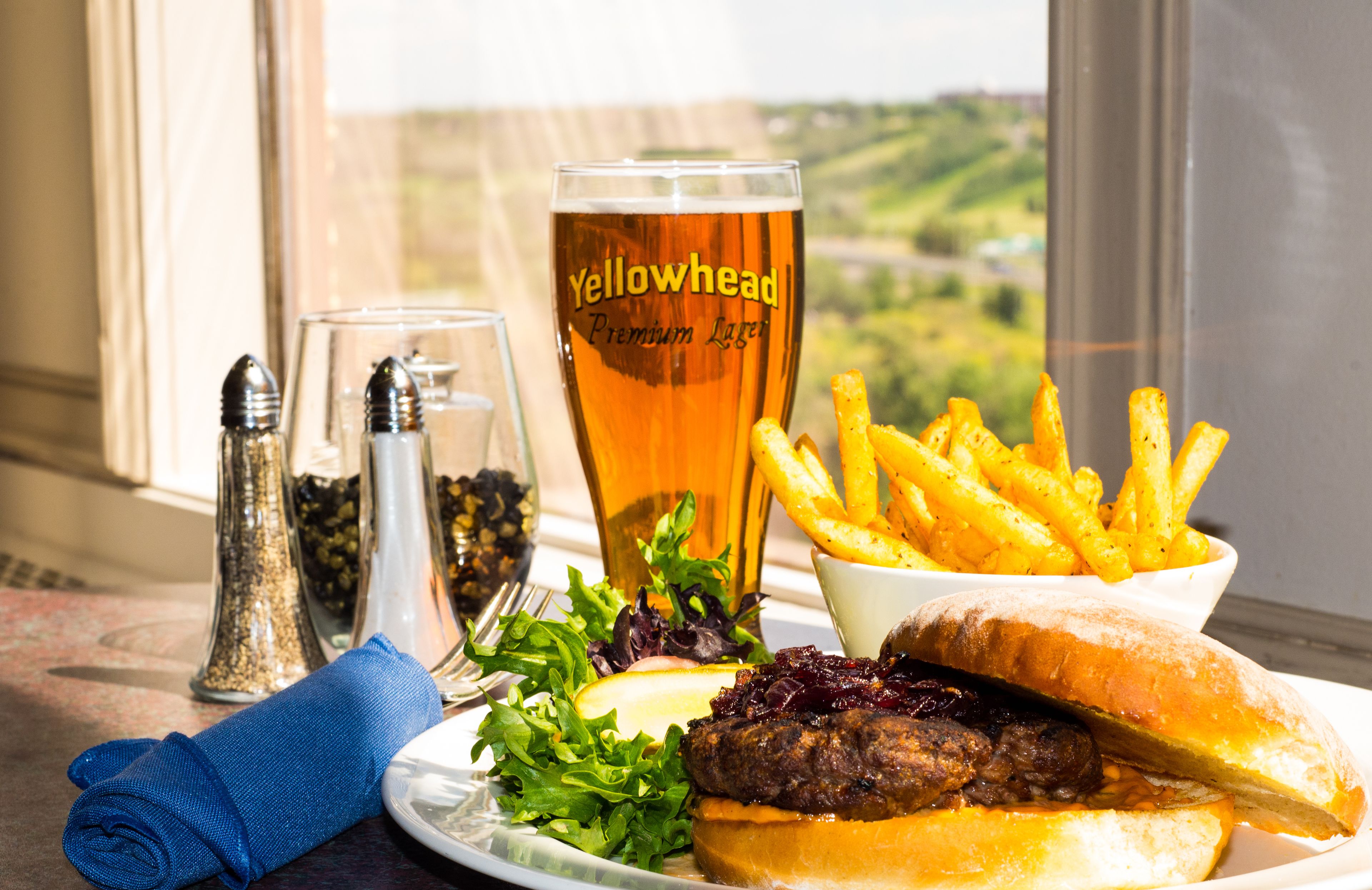 A meal with a burger, fries, and a glass of beer on a table with a window view.