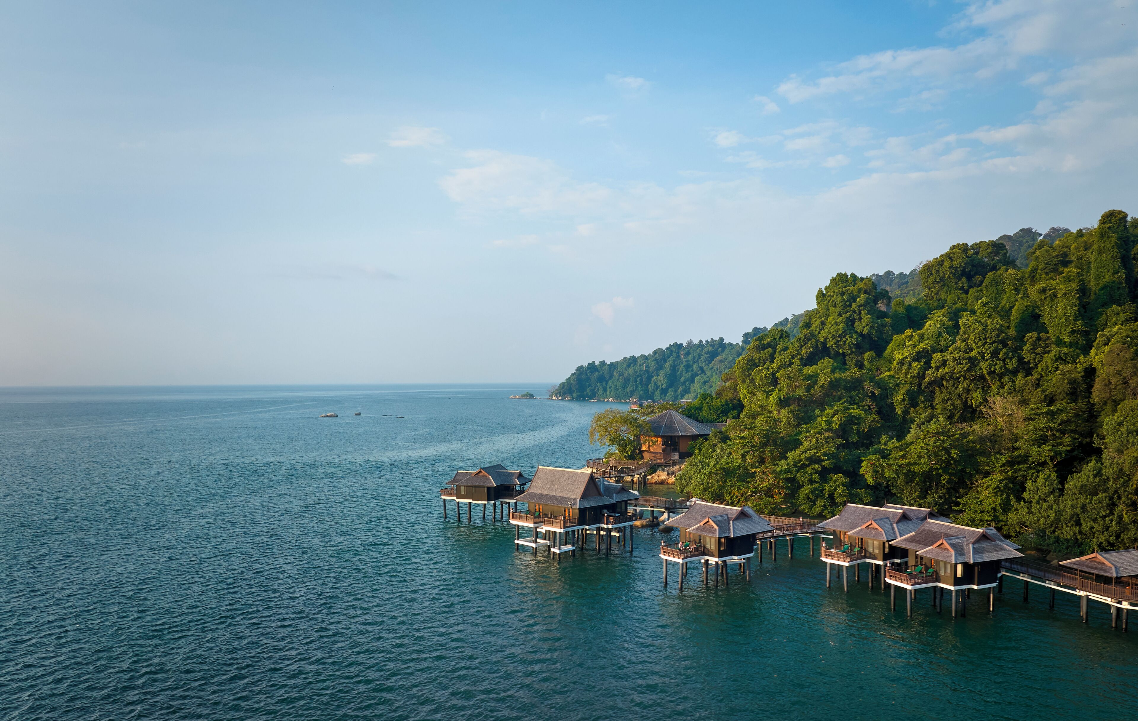 Overwater bungalows on stilts with a view of the sea and a forested hillside.