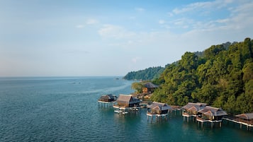 Overwater bungalows on stilts with a view of the sea and a forested hillside.