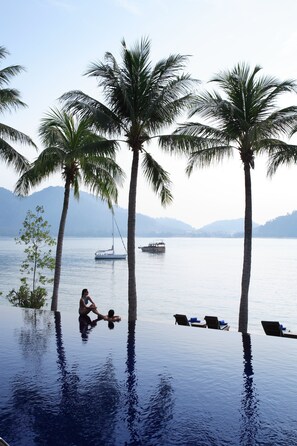 An infinity pool with palm trees and lounge chairs, overlooking a calm lake with boats and mountains in the background.