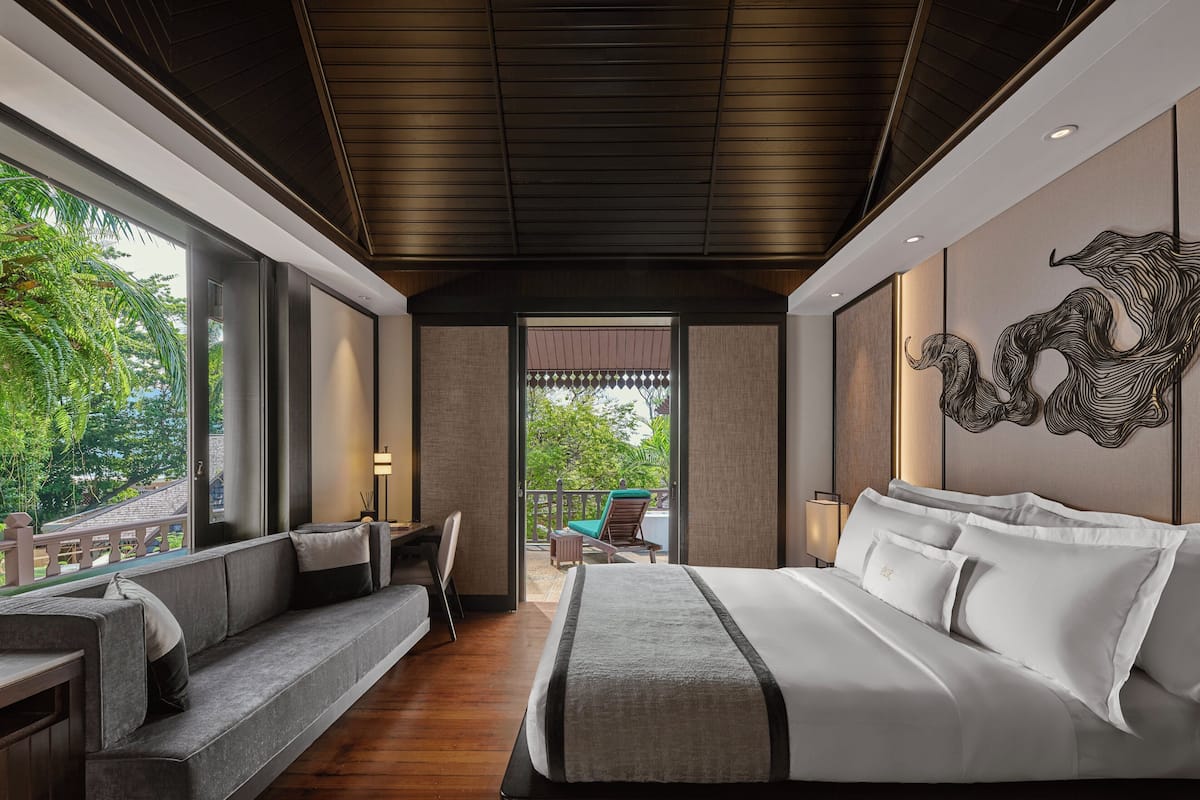 A modern hotel room with a large bed, a seating area with a sofa and chairs, a view of the outdoors, and a decorative wall piece.