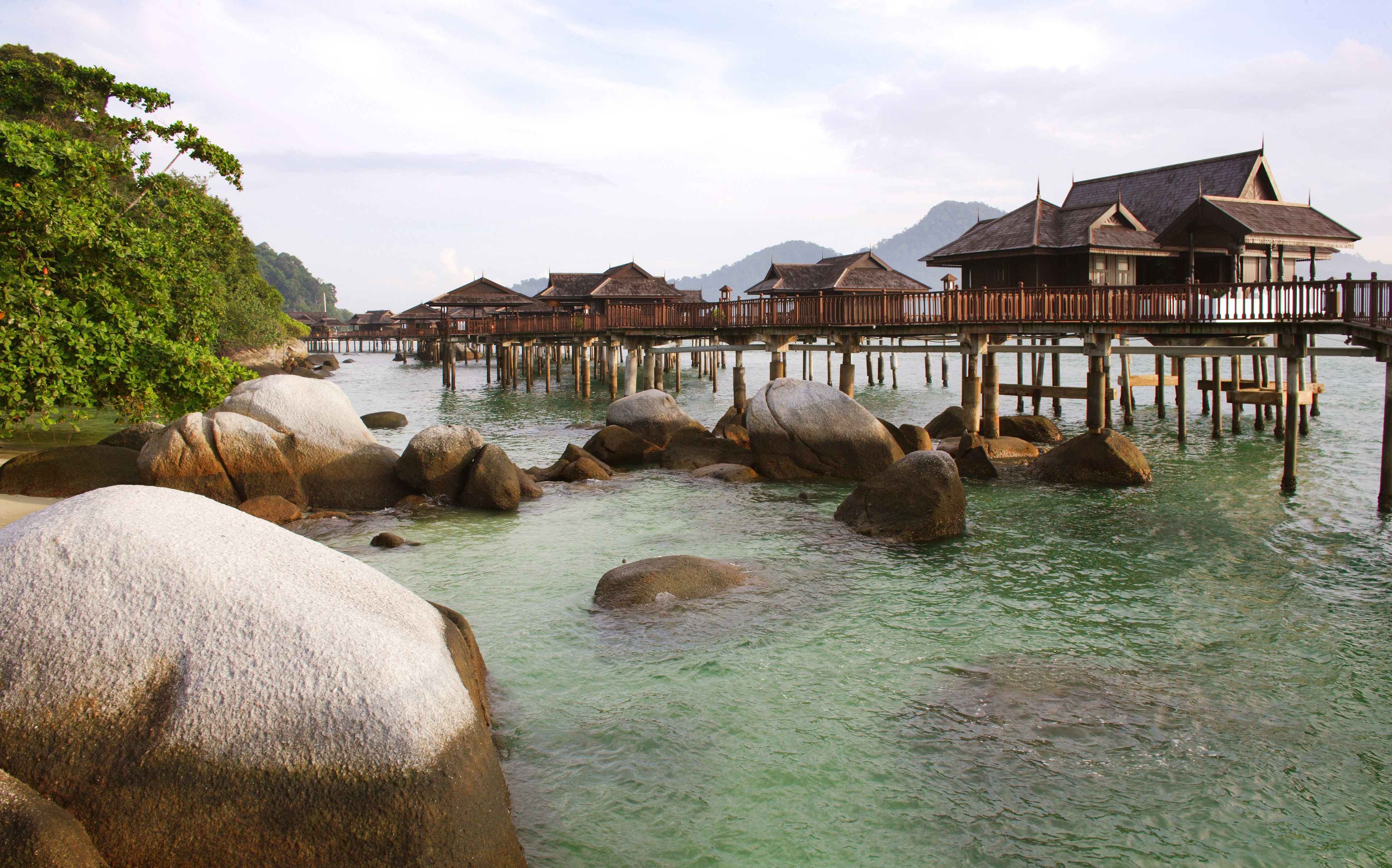 Overwater bungalows with wooden structures and thatched roofs, supported by pillars in clear water, surrounded by large rocks and greenery.