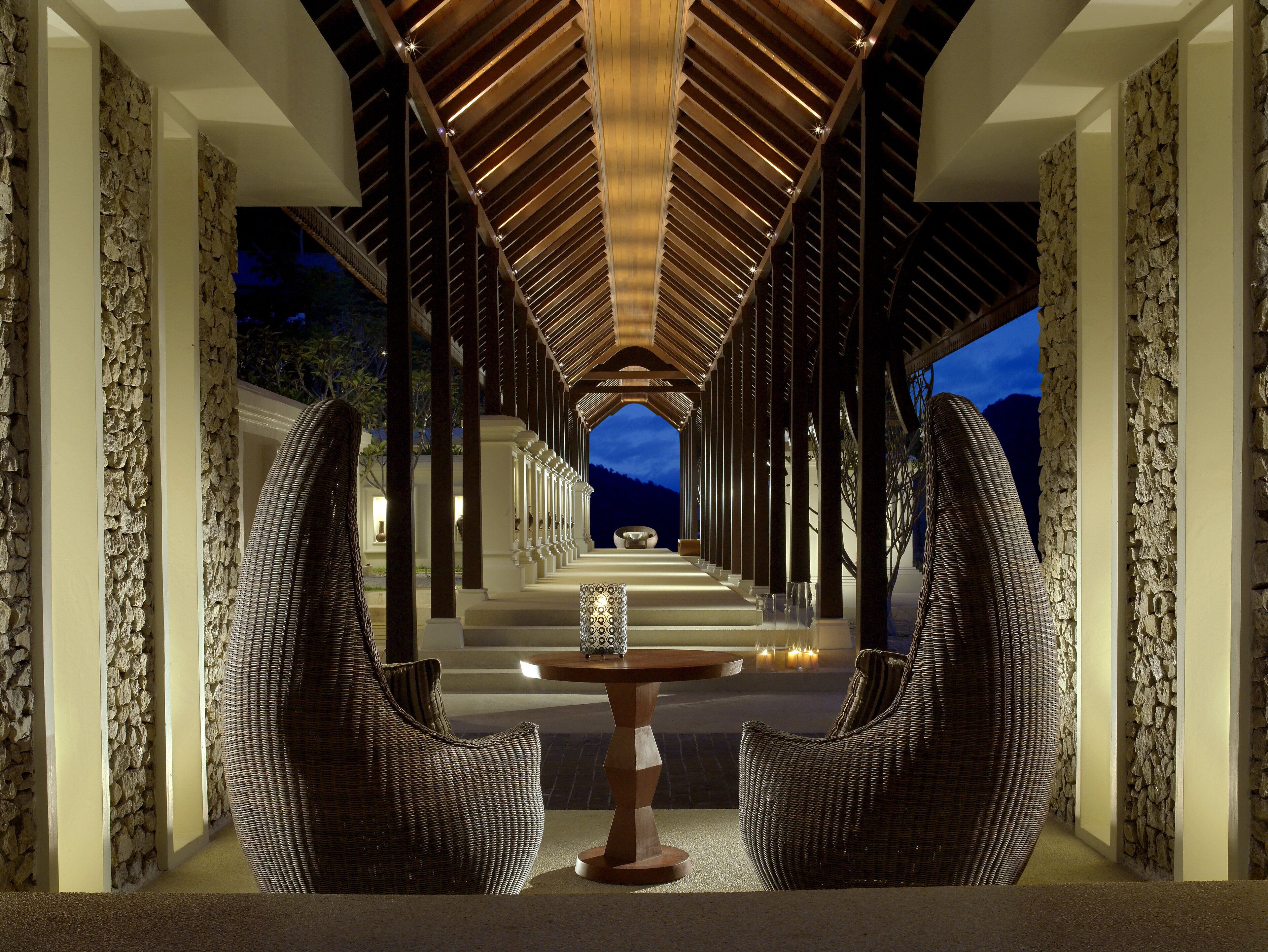 A corridor with a wooden ceiling and stone walls, leading to a seating area with two wicker chairs and a table.