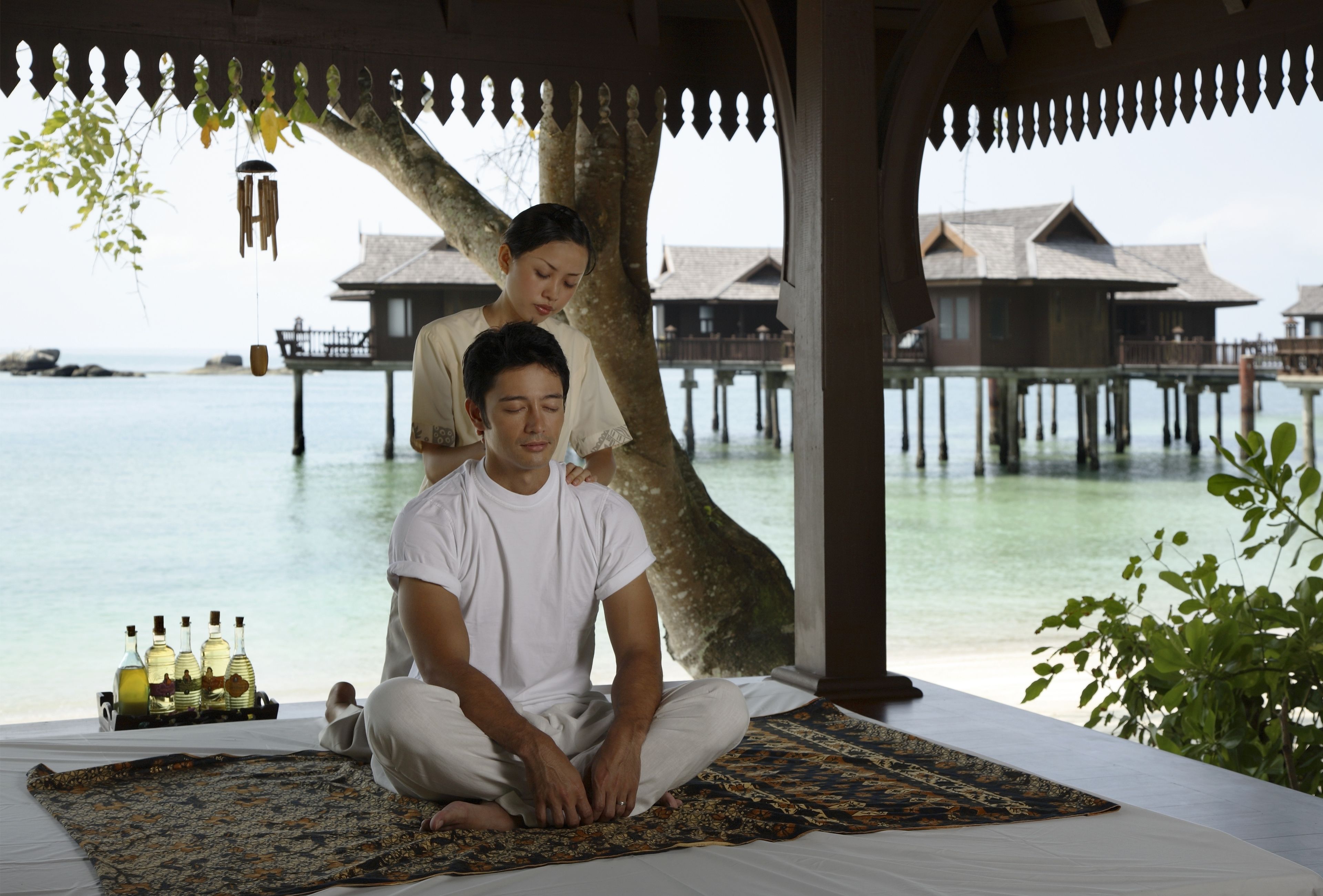 A man and a woman sitting on a bed under a tree, with a view of overwater bungalows and the sea.
