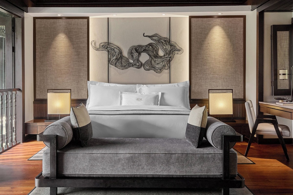 A modern hotel room with a large bed, two bedside lamps, a desk with a chair, and a decorative wall panel.