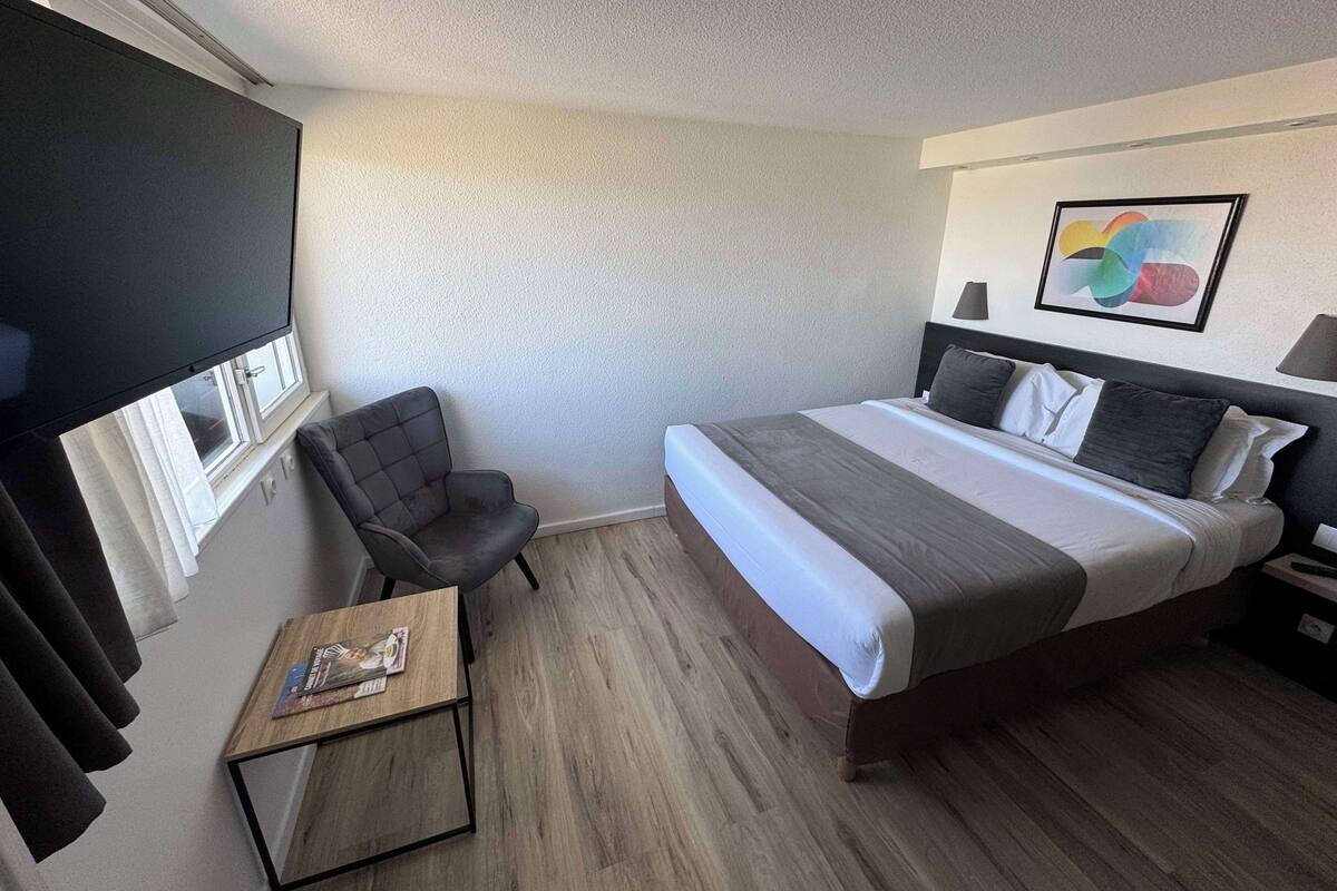 A hotel room with a bed, a chair, a small table, and a window with curtains.