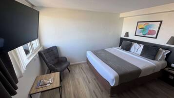 Quarto de hotel com uma cama, uma cadeira, uma mesinha e uma janela com cortinas.