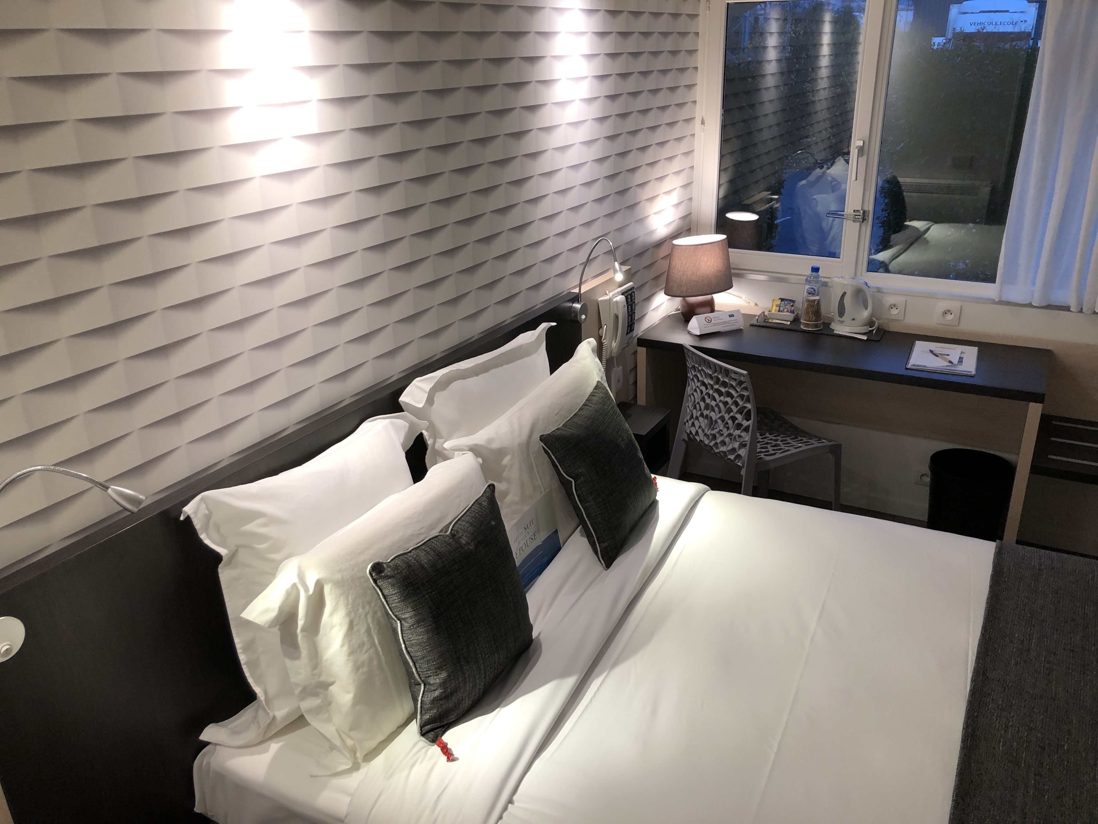 Quarto de hotel com cama, almofadas, escrivaninha, cadeira, abajur, telefone e janela.