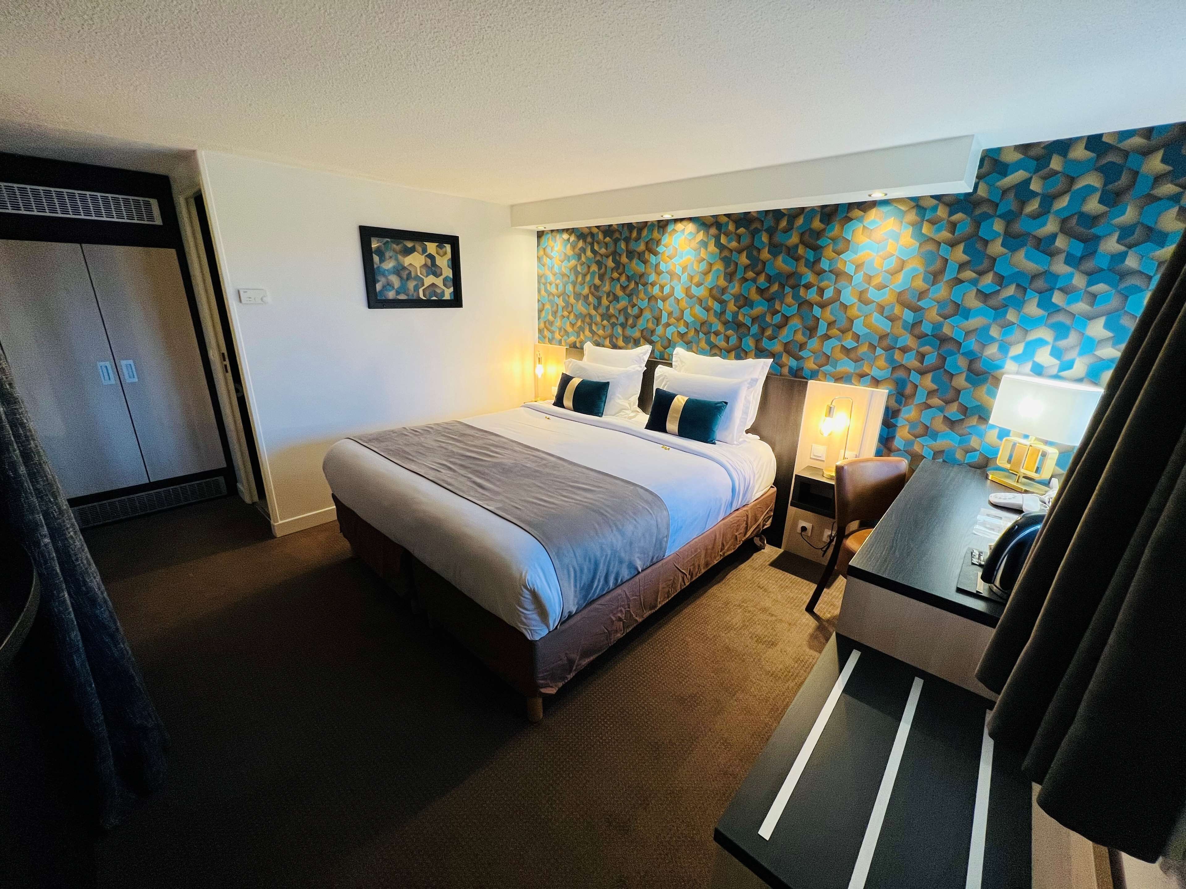 A hotel room with a large bed, a desk with a lamp, and a wardrobe.