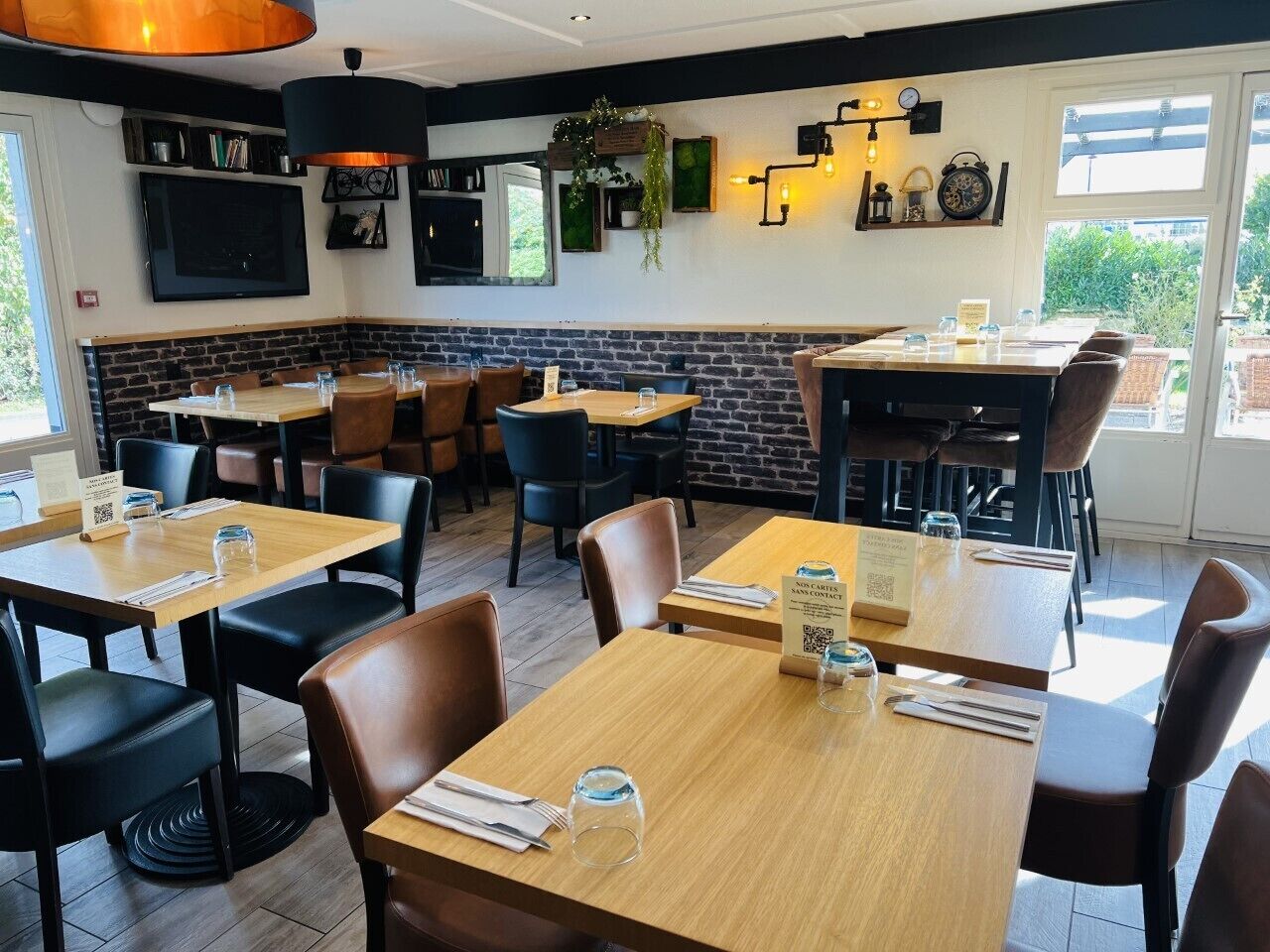 A restaurant with wooden tables and chairs, a brick accent wall, and a large window providing a view of the outdoors.
