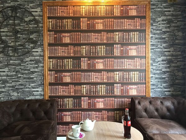 A cozy reading area with leather armchairs, a wooden table with a teapot and a bottle, and a bookshelf filled with books against a stone wall.