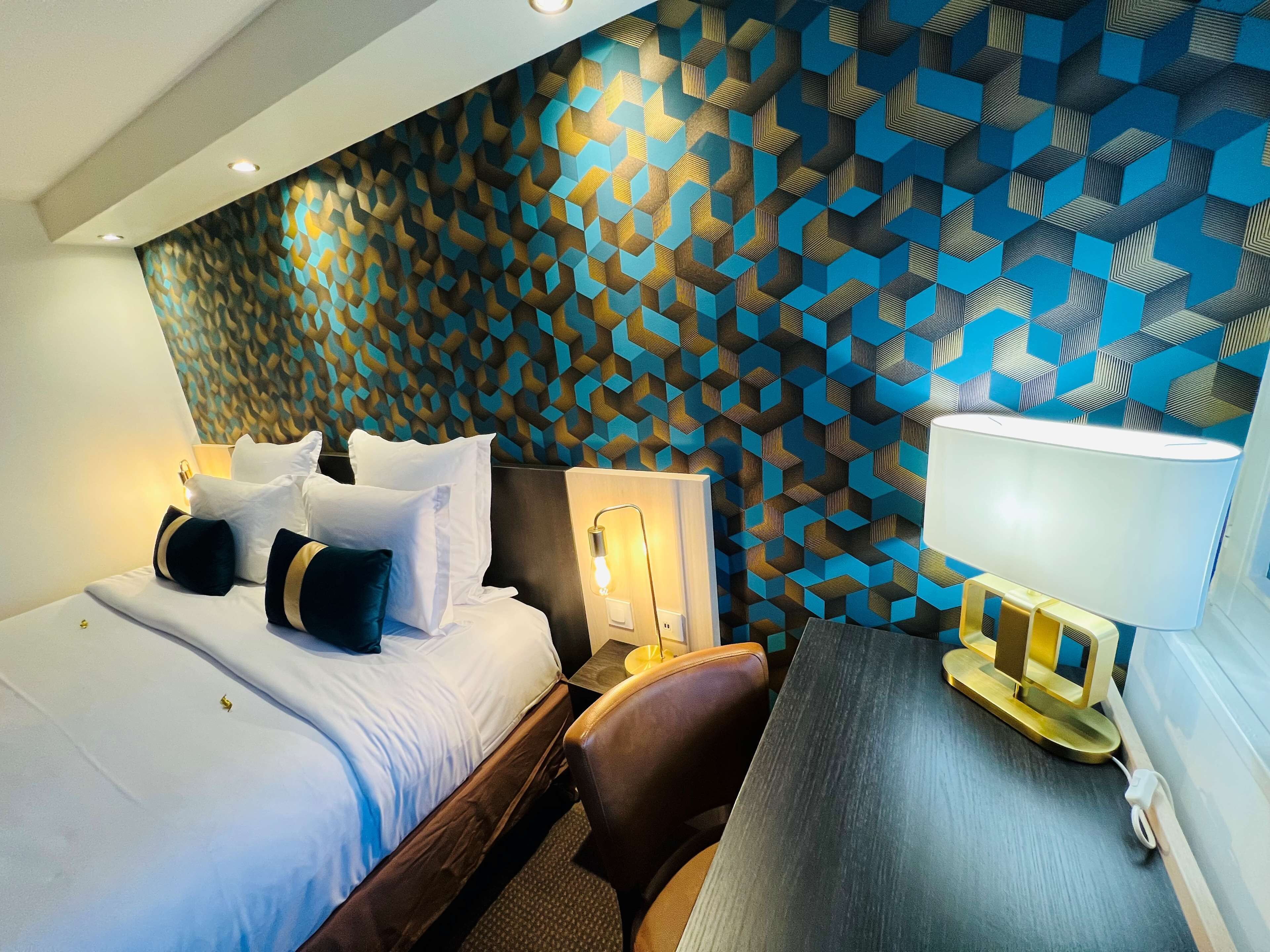 A hotel room with a modern design, featuring a geometric patterned wall, a neatly made bed with multiple pillows, a desk with a lamp, and a chair.
