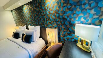 A hotel room with a modern design, featuring a geometric patterned wall, a neatly made bed with multiple pillows, a desk with a lamp, and a chair.