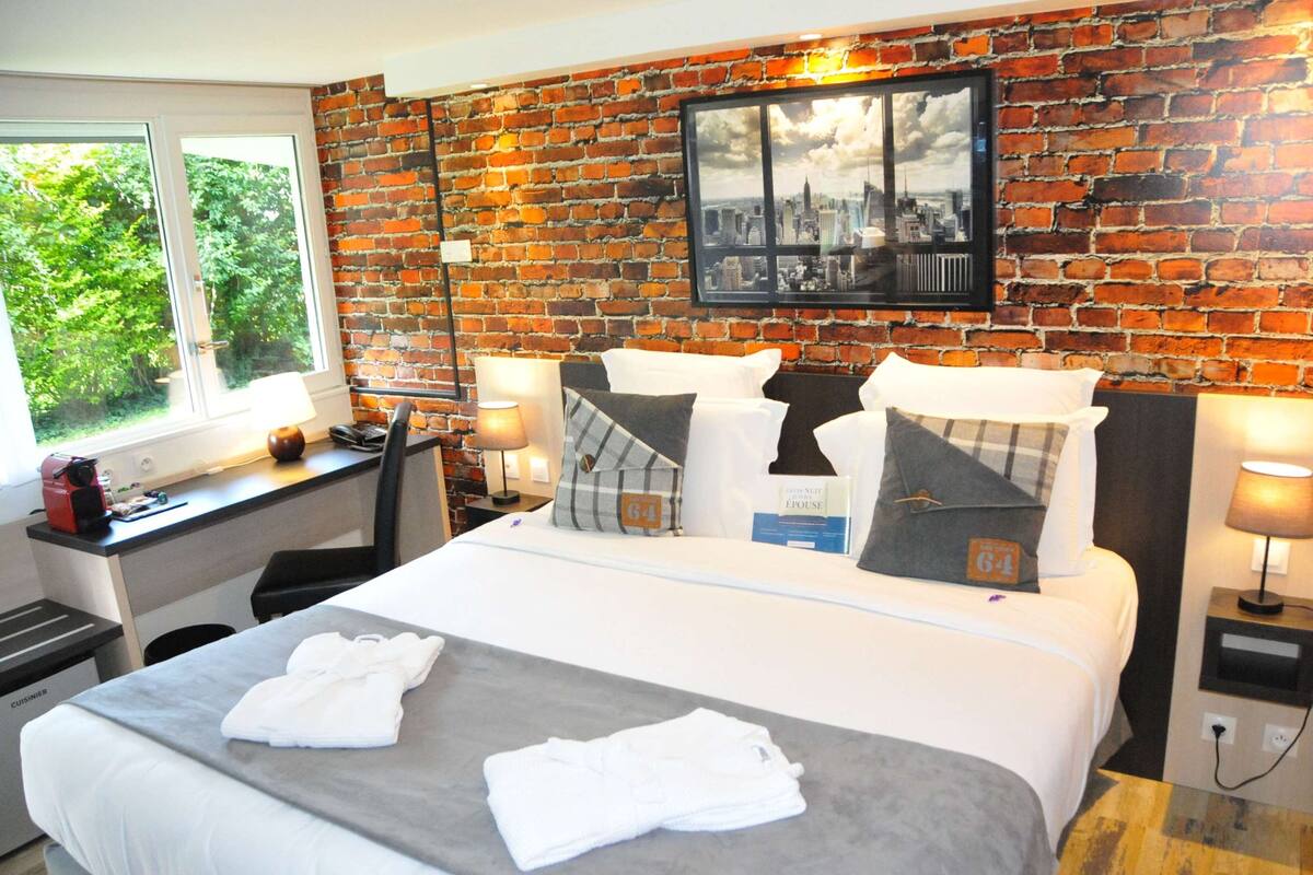 A hotel room with a brick wall, a bed with white bedding, a desk with a lamp, and a window with a view of greenery.