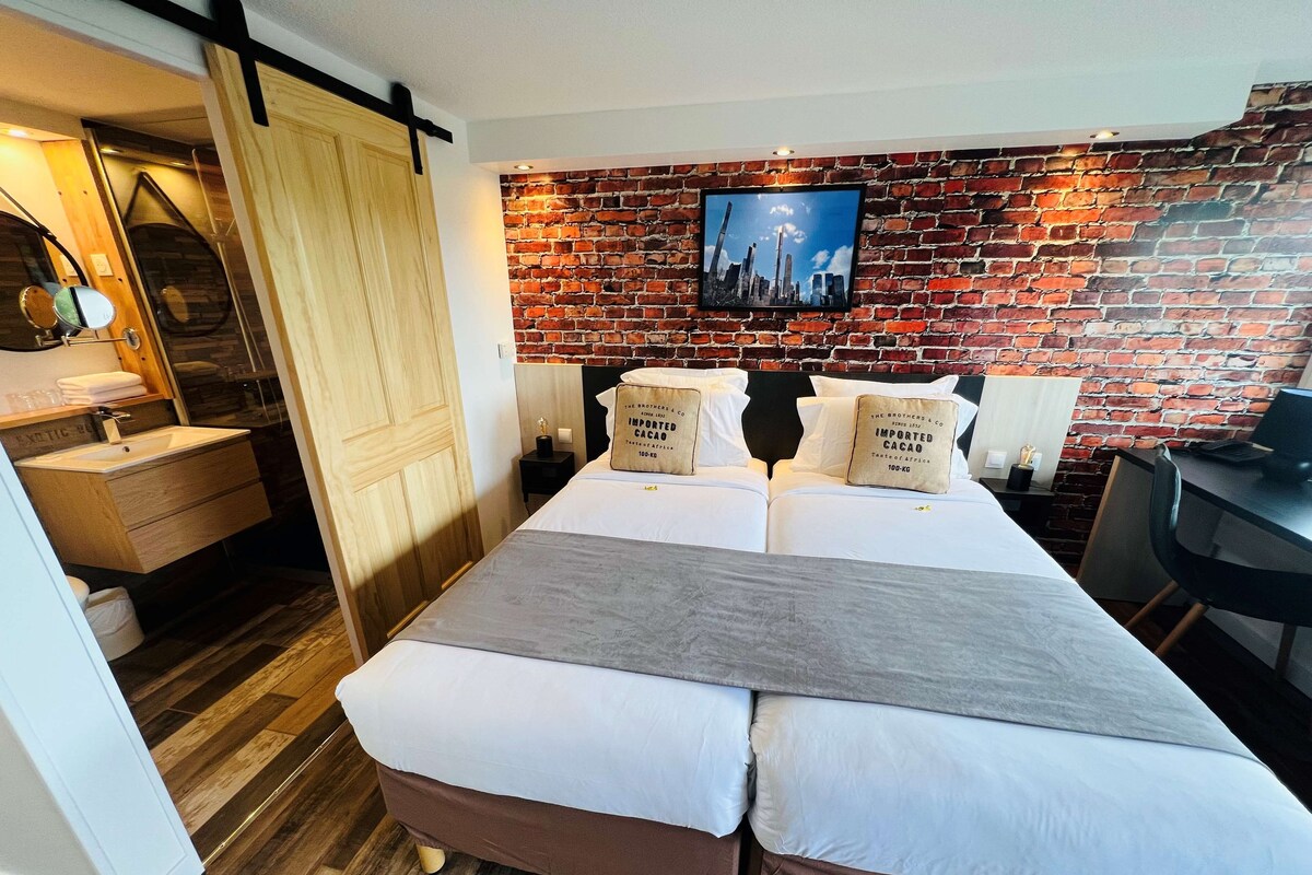 A hotel room with a brick wall, a bed with two pillows, a desk, and a bathroom with a wooden cabinet and mirror.