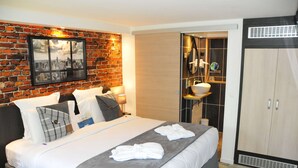 A hotel room with a brick wall, a bed with white bedding, a wardrobe, and a bathroom with a mirror and shelves. - Kyriad Lisieux (Lisieux)