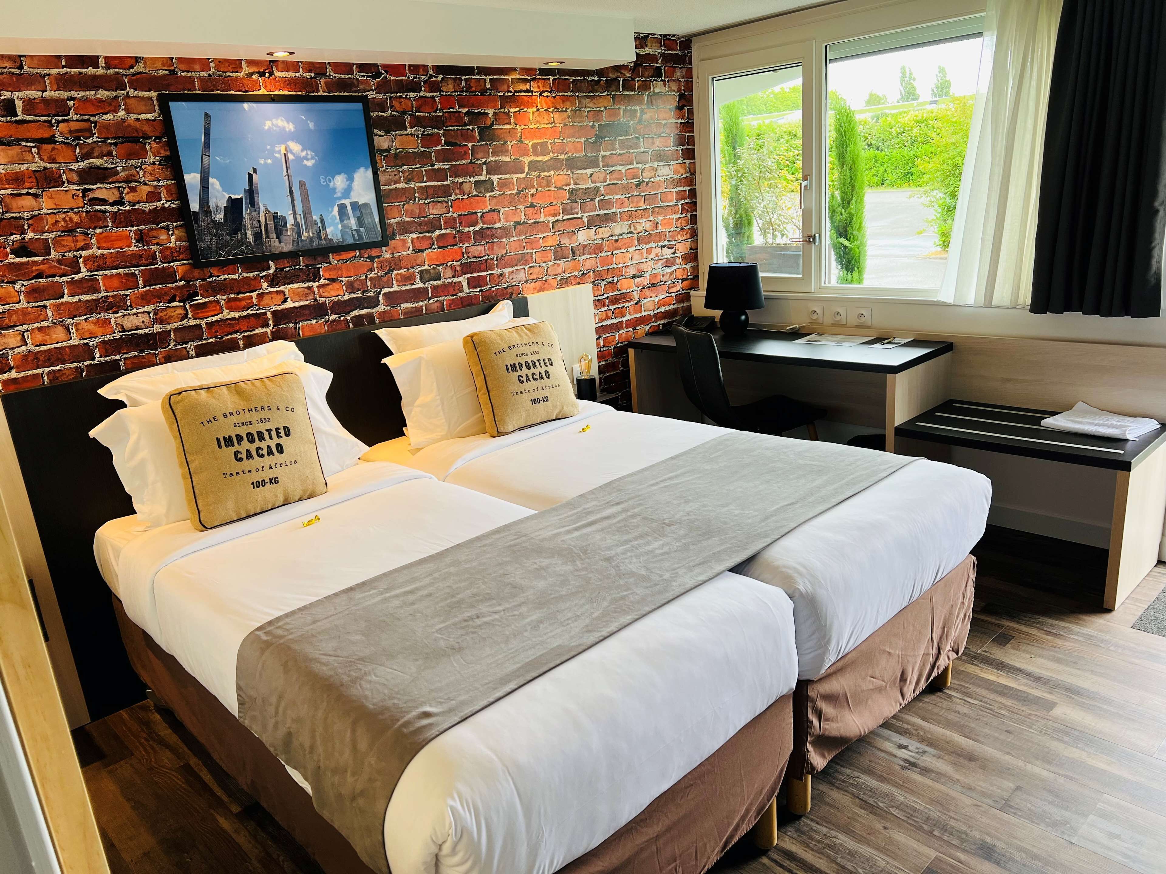 A hotel room with a brick wall, a bed with white linens, two pillows with text, a desk with a chair, and a window with curtains.