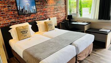 A hotel room with a brick wall, a bed with white linens, two pillows with text, a desk with a chair, and a window with curtains.
