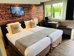 A hotel room with a brick wall, a bed with white linens, two pillows with text, a desk with a chair, and a window with curtains. - Kyriad Lisieux (Lisieux)