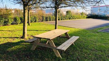 A picnic table in a grassy area with trees and a parking lot in the background.