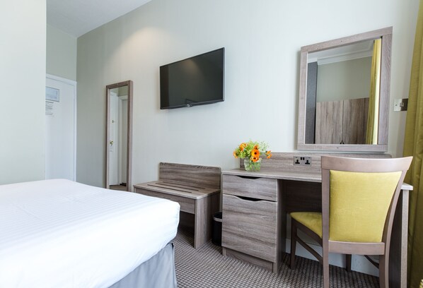 Habitación de hotel con cama, escritorio, silla, televisión, espejo y un jarrón con flores.