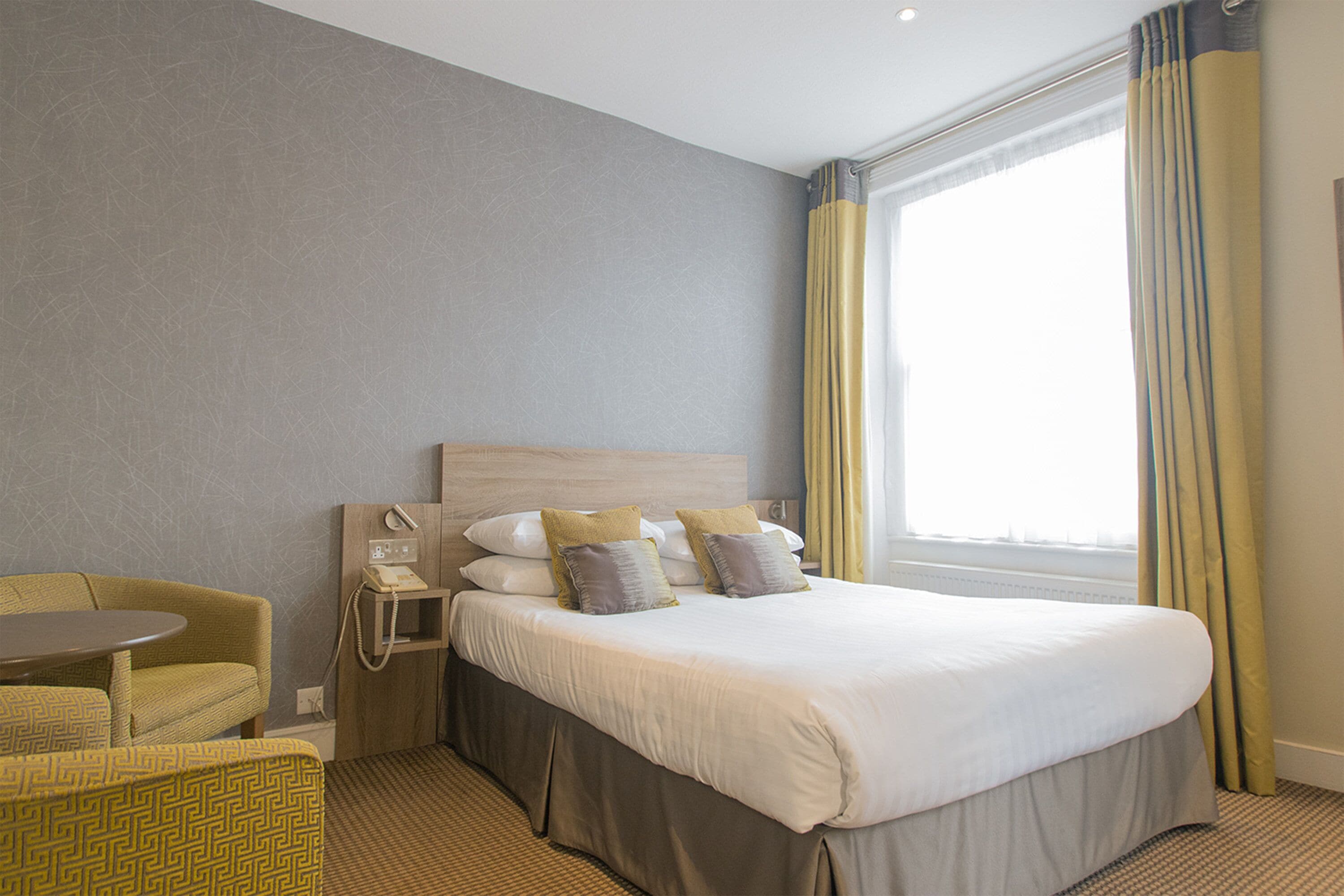 Quarto de hotel com cama, uma cadeira, mesa de cabeceira e janela com cortinas.