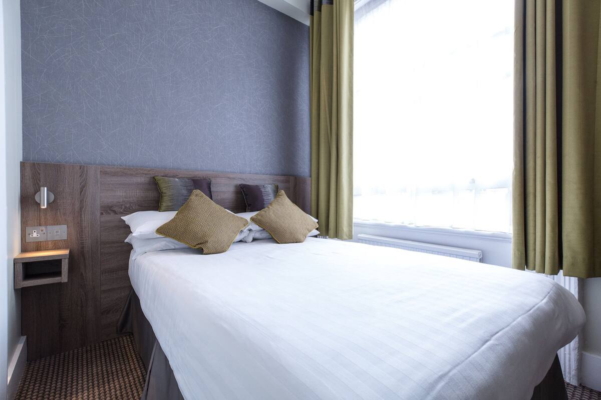 Une chambre d’hôtel avec un lit bien fait, une tête de lit en bois, une petite table de chevet et une fenêtre avec des rideaux.