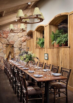 A long dining table set for a meal, surrounded by wooden chairs, with a stone wall and wooden paneling in the background.