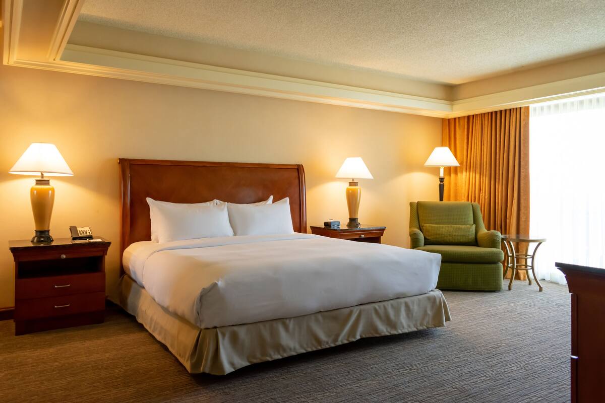 A hotel room with a large bed, two bedside tables with lamps, a green armchair, and a small table.