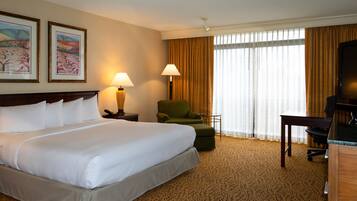 A hotel room with a large bed, two bedside lamps, a green armchair, a desk with a TV, and a window with curtains.