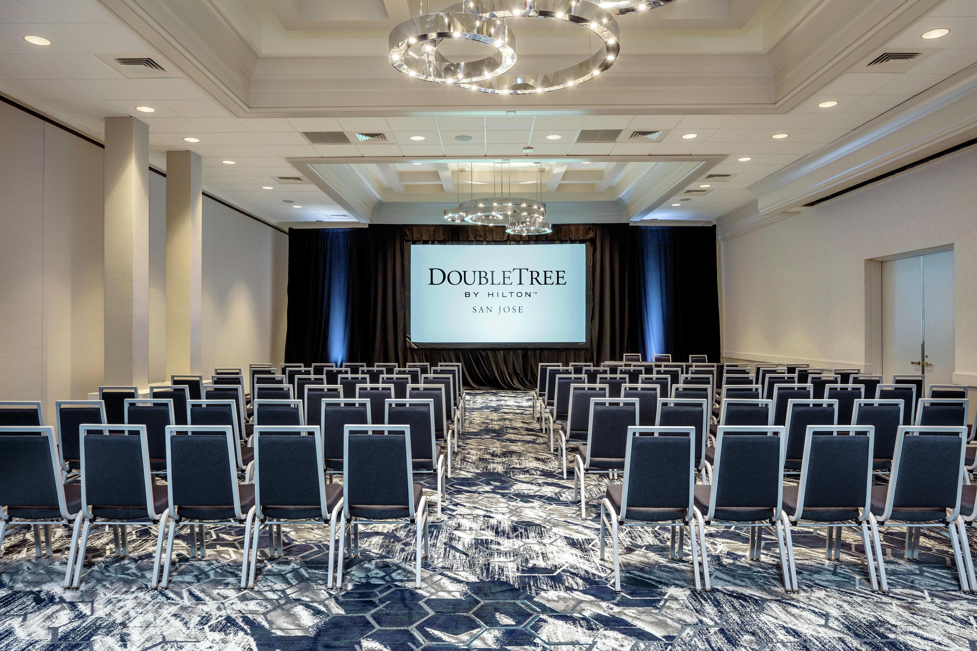 Une salle de conférence avec des rangées de chaises, un grand écran affichant le texte « DOUBLETREE BY HILTON SAN JOSE », et un tapis à motifs.