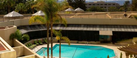 A swimming pool with lounge chairs and umbrellas, surrounded by palm trees and a building with a curved roof.