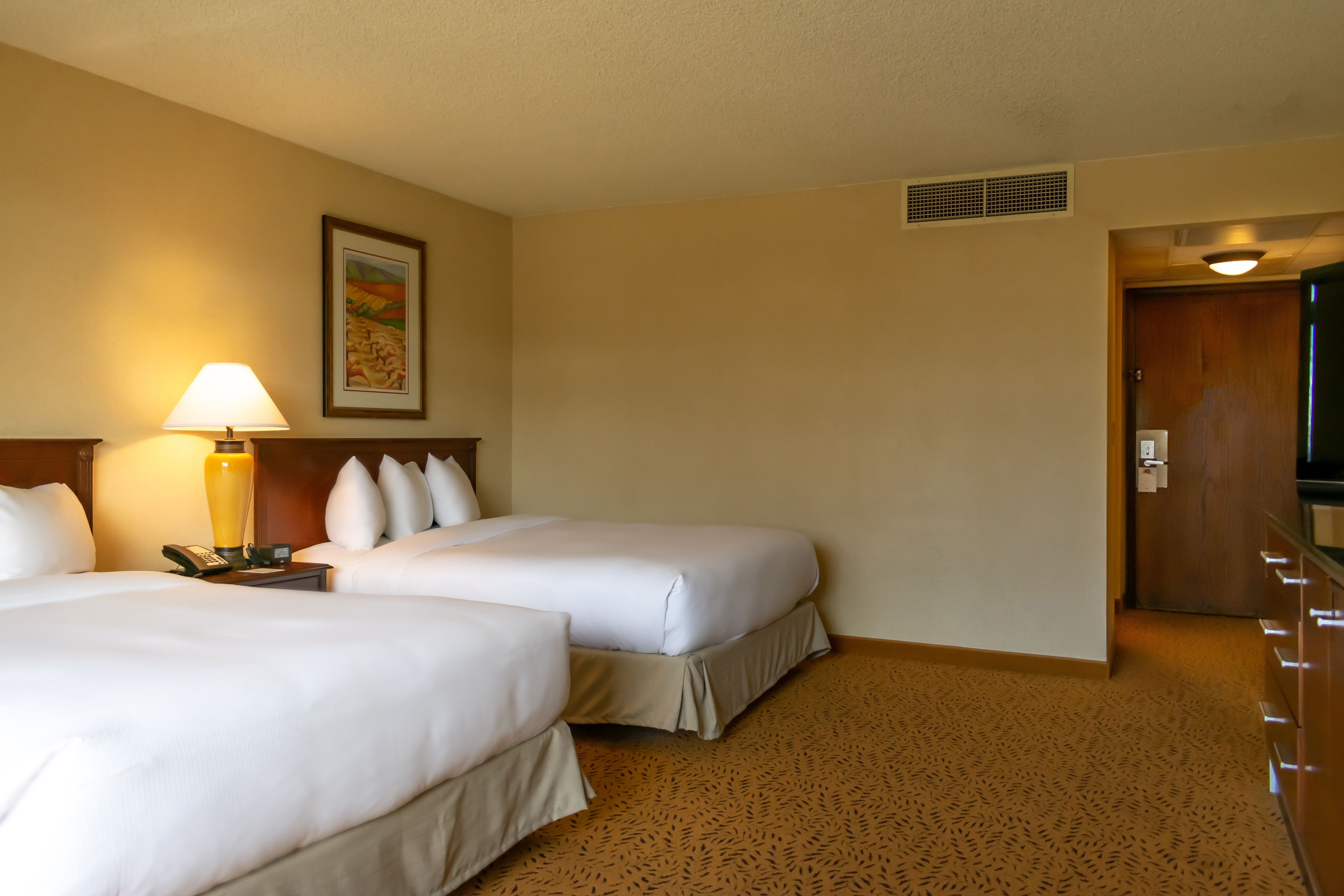 Habitación de hotel con dos camas, una mesita de noche con lámpara, un cuadro en la pared y una puerta que da a otra habitación.