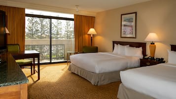 A hotel room with two beds, a desk, a chair, a small table, and a view of trees through the window.