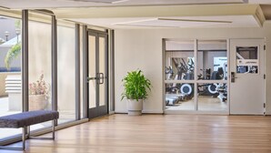 Moderni hotellin aula, jossa on puulattia, penkki, ruukussa oleva kasvi ja näkymä kuntosalille lasiovien läpi.