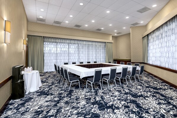 Une salle de conférence dotée d’une grande table rectangulaire entourée de chaises, d’un sol recouvert de moquette et d’une grande fenêtre avec des rideaux.