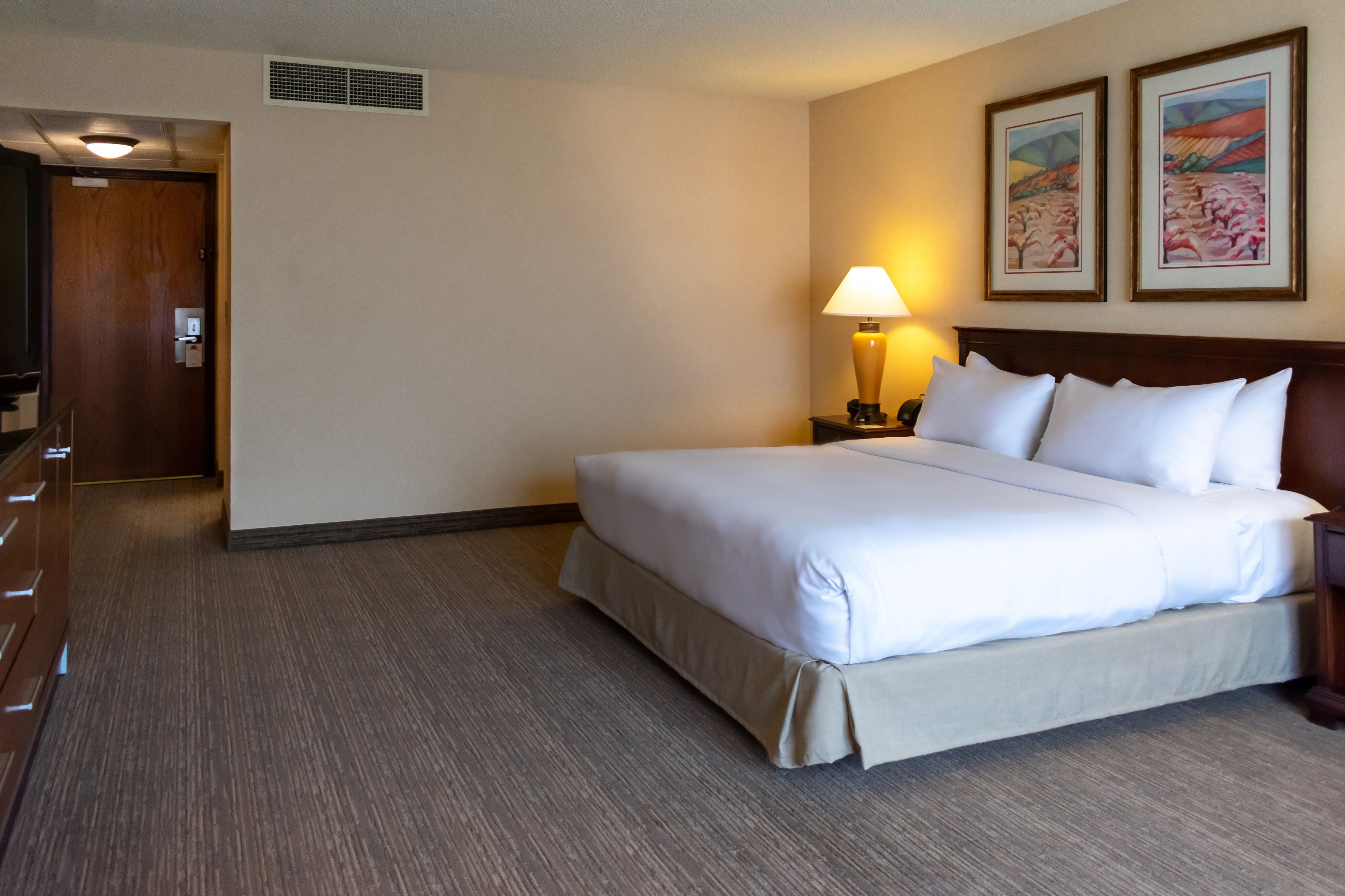 Une chambre d’hôtel avec un grand lit, une table de chevet avec une lampe, une tête de lit en bois et deux tableaux encadrés au mur.