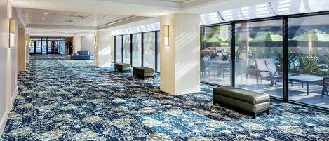 A hotel corridor with patterned carpet, seating areas, and large windows offering views of outdoor seating.