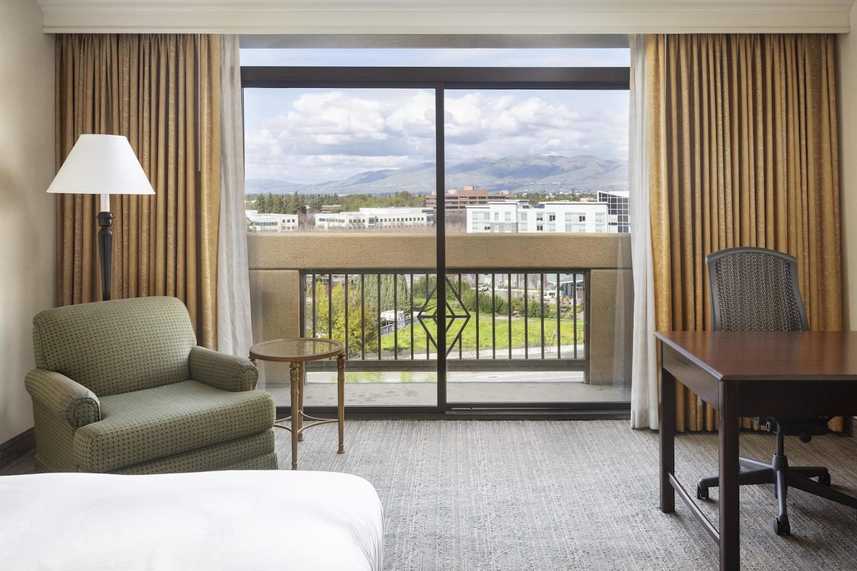 A hotel room with a bed, a chair, a table, a lamp, and a view of buildings and mountains through a large window.