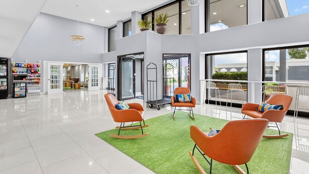 A spacious lobby with modern orange rocking chairs, a green artificial turf area, and large windows offering views of the outside.