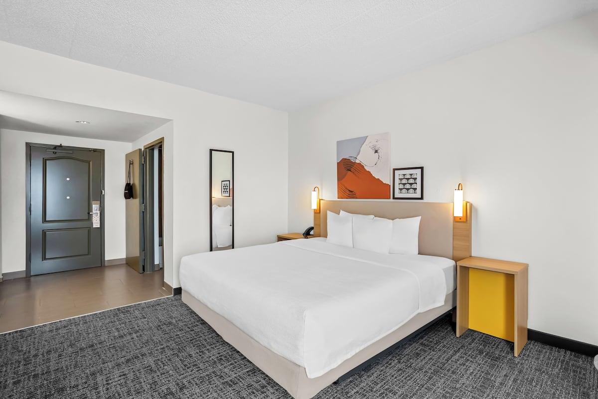 Une chambre d’hôtel avec un grand lit, des tables de chevet, une petite table, une lampe fixée au mur et une porte donnant sur une autre pièce.