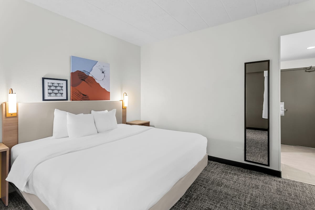 Une chambre d’hôtel avec un grand lit, deux tables de chevet, une commode, une lampe fixée au mur, un tableau encadré et un miroir.