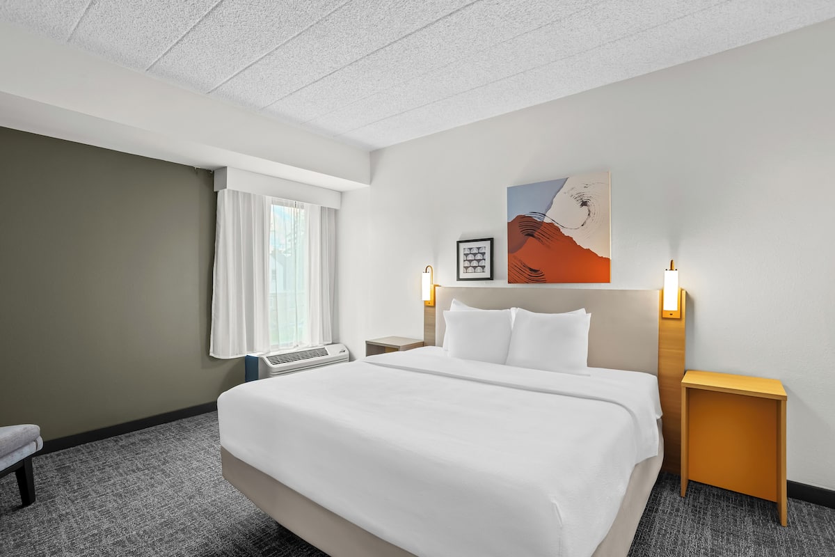 Une chambre d’hôtel avec un grand lit, une table de chevet, une commode, une fenêtre avec des rideaux, une œuvre d’art accrochée au mur et un luminaire au plafond.
