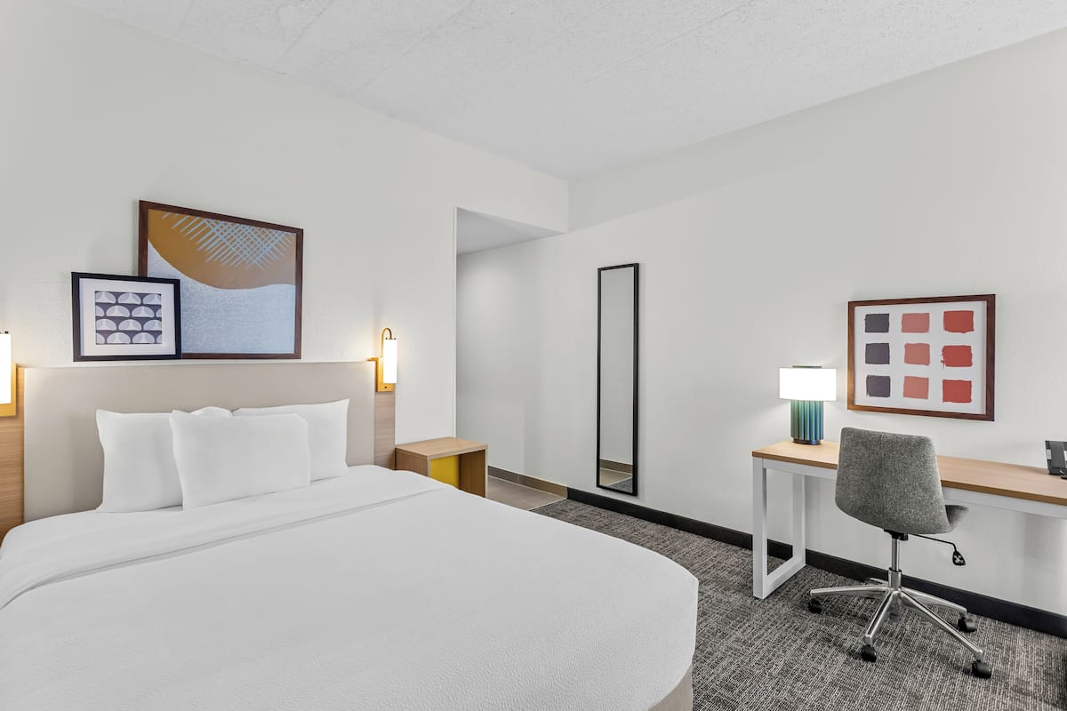 Une chambre d’hôtel avec un grand lit, un bureau, une chaise, un miroir et des œuvres d’art accrochées aux murs.