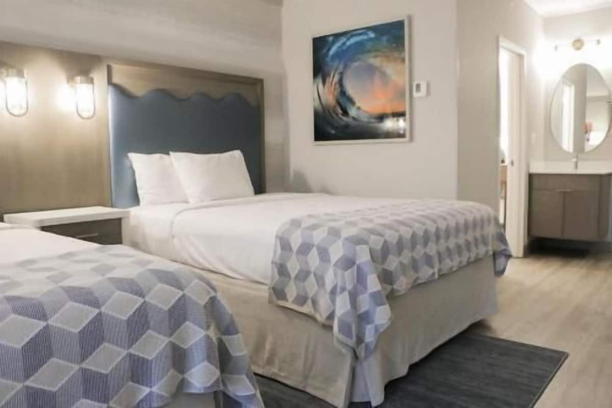A hotel room with two beds, a painting on the wall, a bathroom with a round mirror, and a small desk.