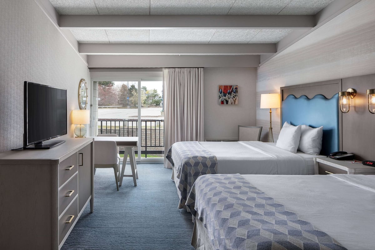 A hotel room with two beds, a desk with a TV, a chair, a window with curtains, and a view of the outside.
