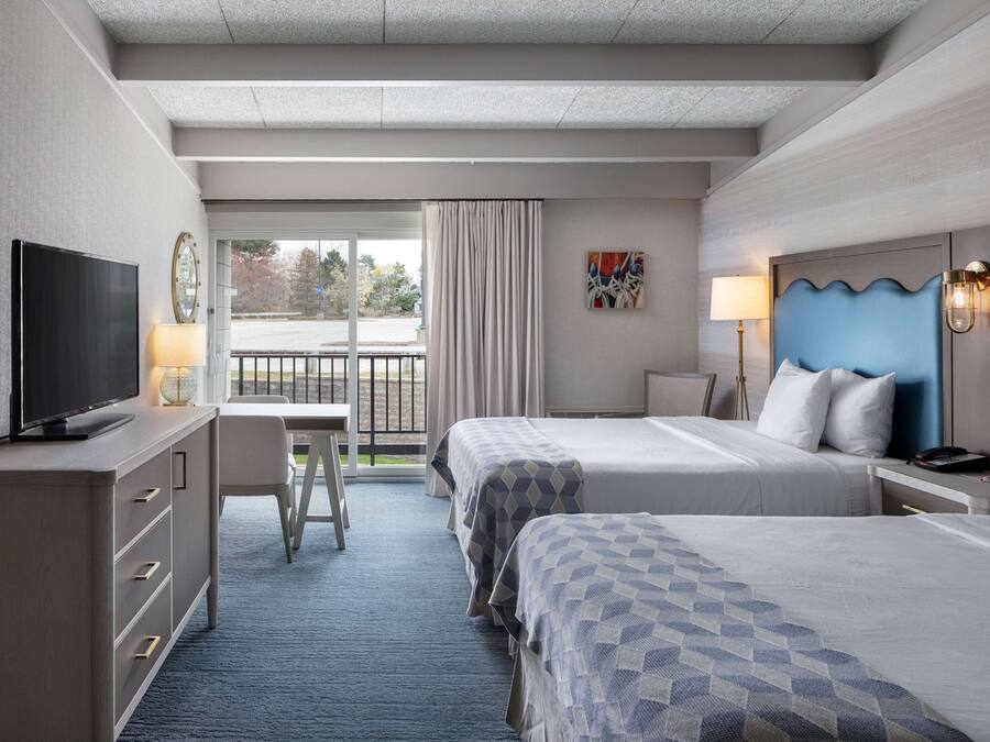 A hotel room with two beds, a desk with a TV, a chair, a window with curtains, and a view of the outside.